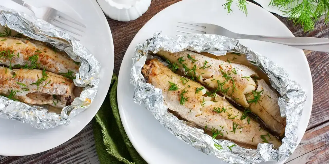 Dwa porcje soczystego mintaja z piekarnika, zapieczonego w folii aluminiowej, posypane świeżym koperkiem.