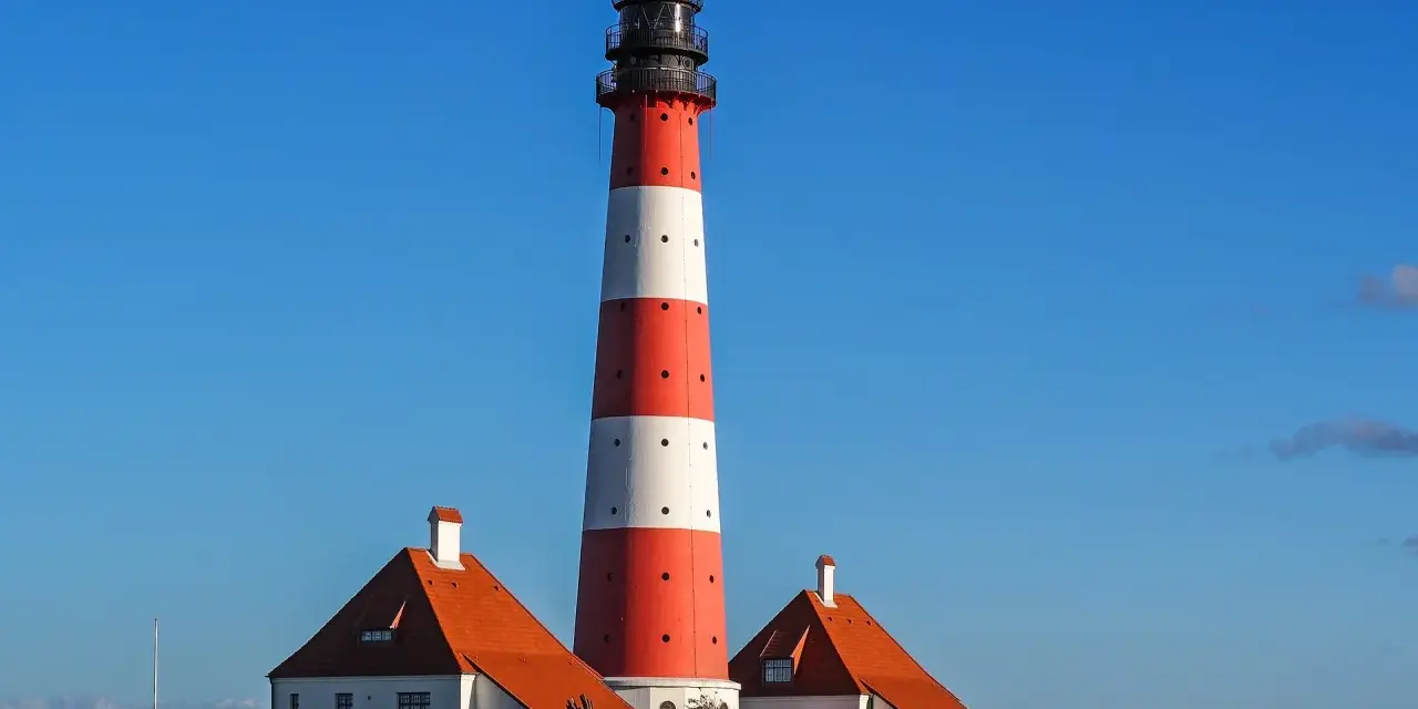 Wysoka, czerwono-biała latarnia morska w Jastarni strzeże wybrzeża. Obok stoją dwa domki z pomarańczowymi dachami.