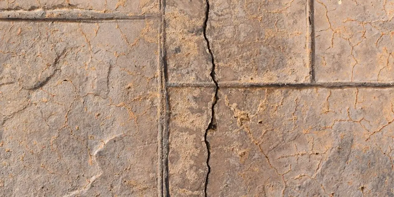 A prominent crack runs through a stamped concrete slab foundation, revealing sand and smaller fissures.