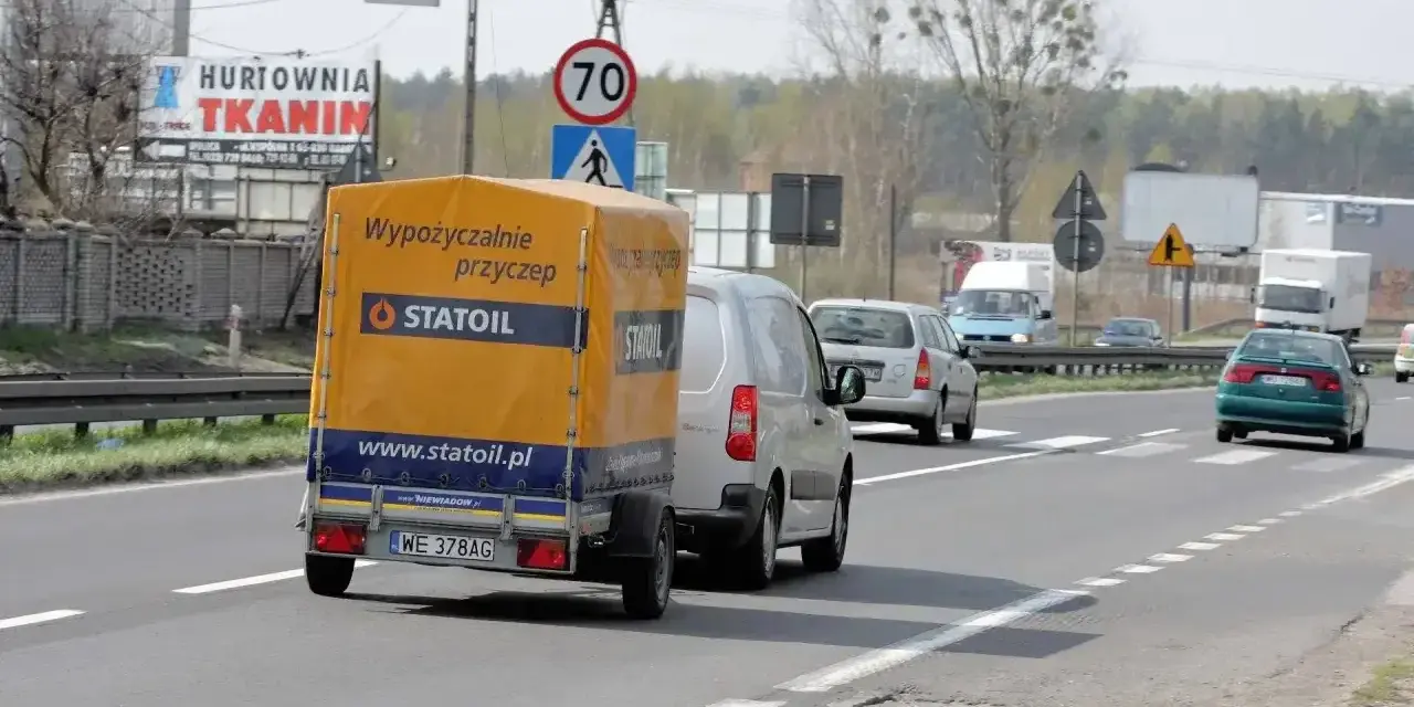 Samochód z żółtą przyczepką Statoil jedzie drogą. Znak ograniczenia do 70 km/h sugeruje, ile można jechać z przyczepką w tym miejscu.