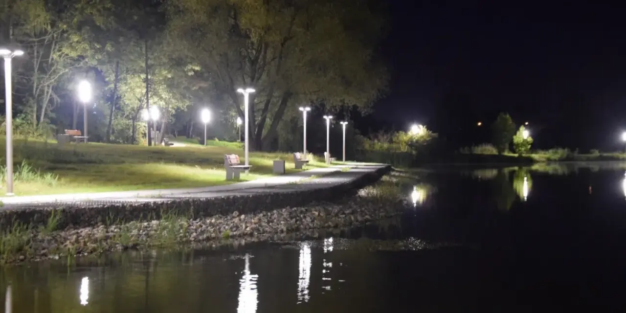Nocny spacer nad zalewem Wolbromskim. Latarnie oświetlają ścieżkę i ławki, odbijając się w spokojnej tafli wody.
