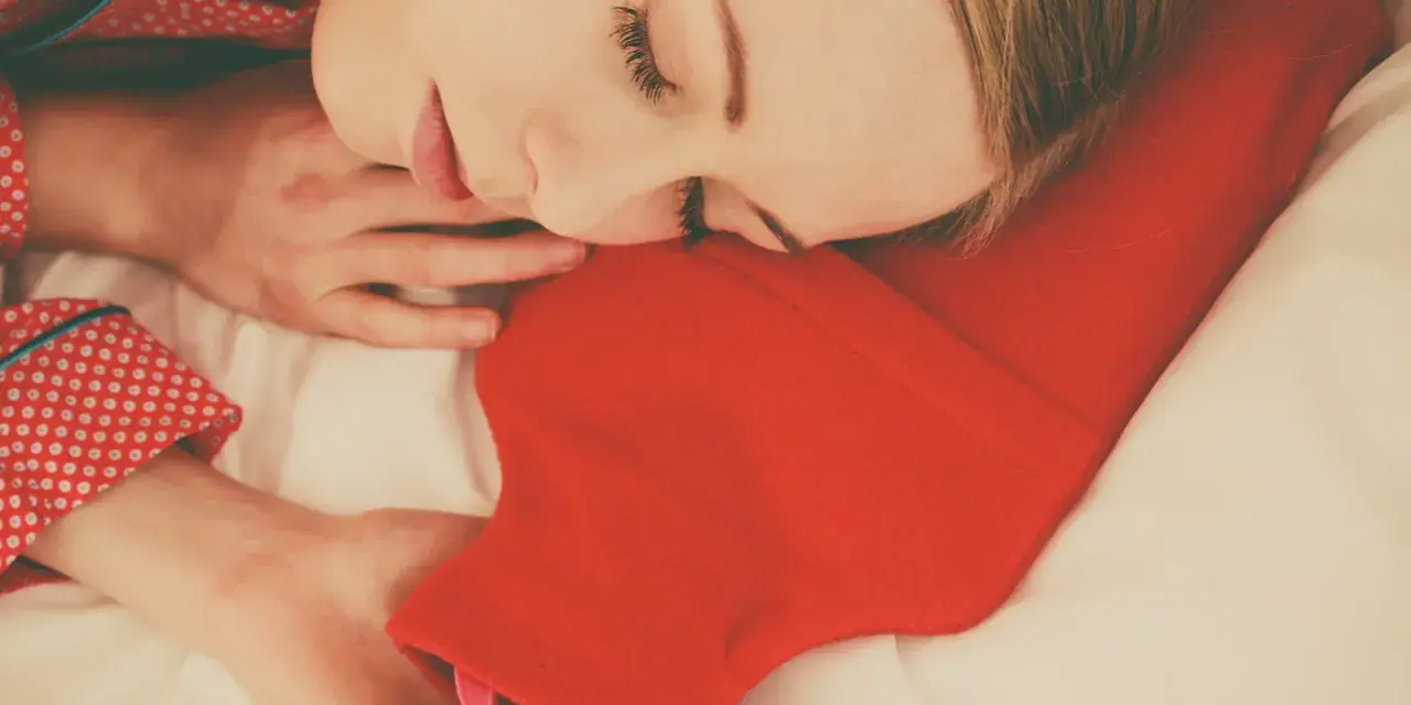 Une femme dort paisiblement, blottie contre une bouillotte rouge. Une astuce pour savoir comment faire une bouillotte avec une serviette pour se réchauffer.