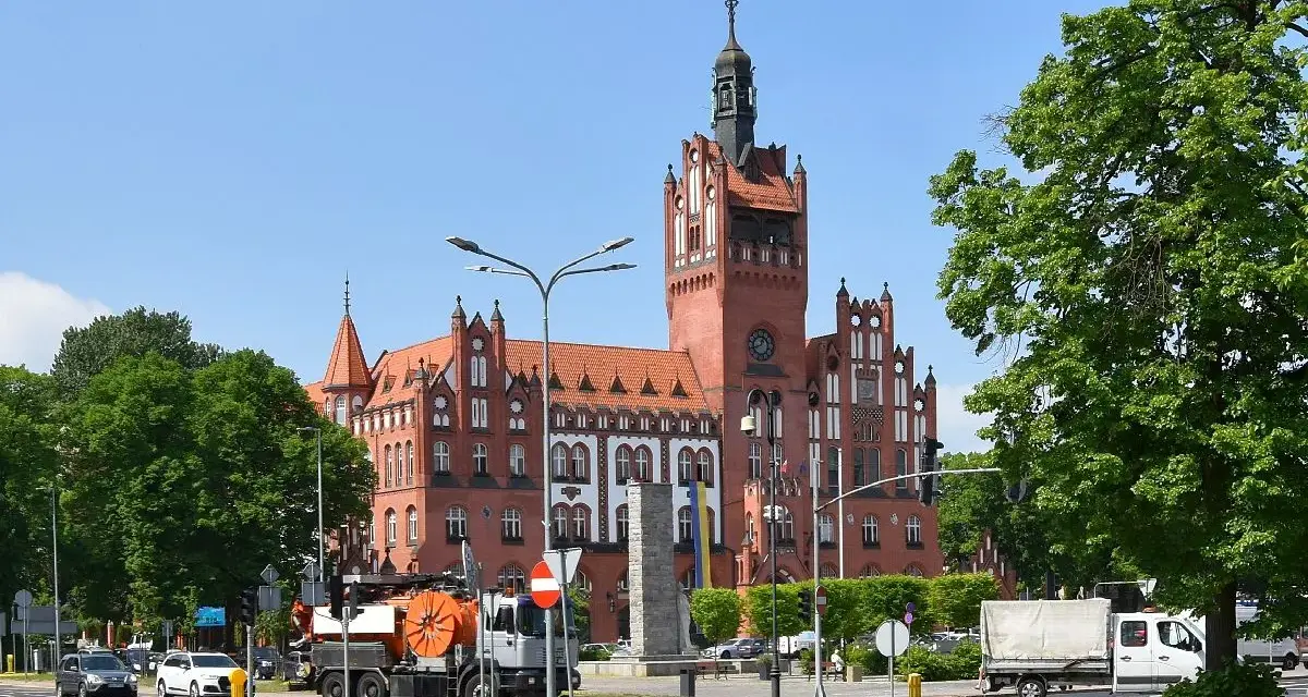 Imponujący ratusz Słupsk z czerwoną cegłą i wieżą góruje nad placem. Wokół drzewa i ruch uliczny.