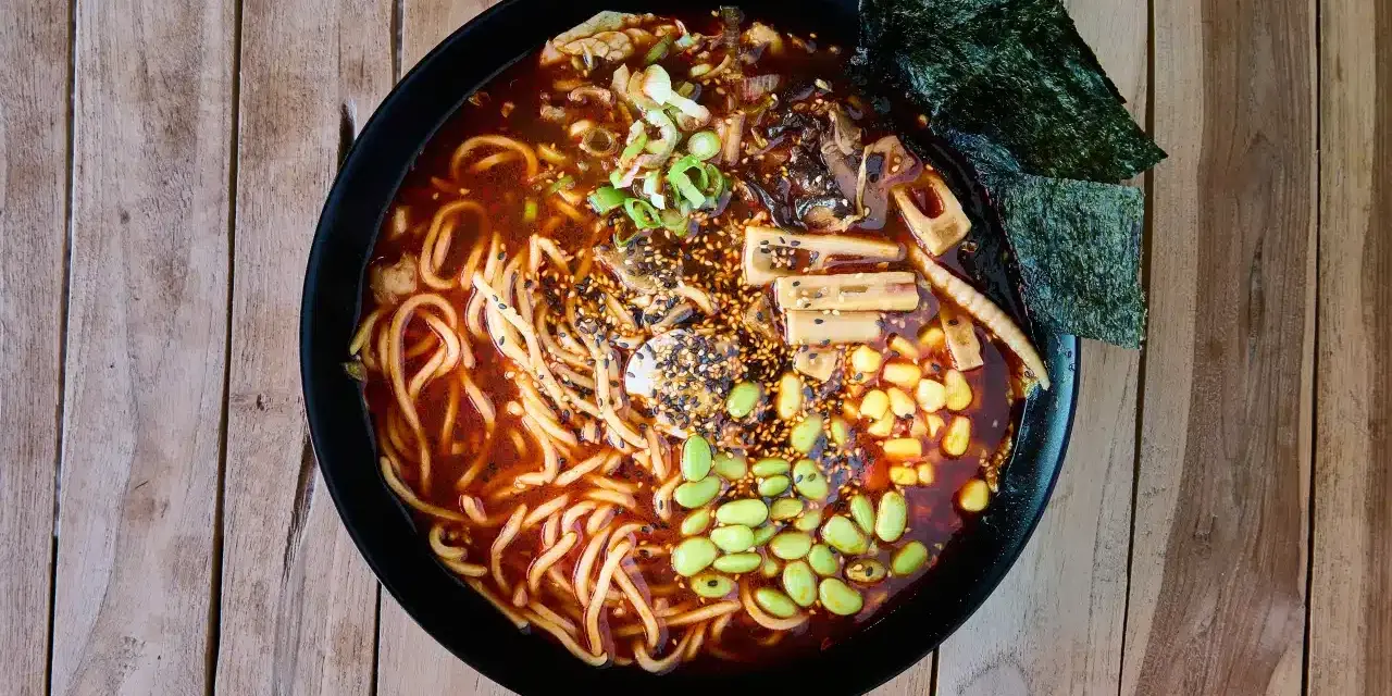 Miseczka aromatycznych dań z makaronem ramen, pełna edamame, kukurydzy, grzybów i nori, na drewnianym tle.