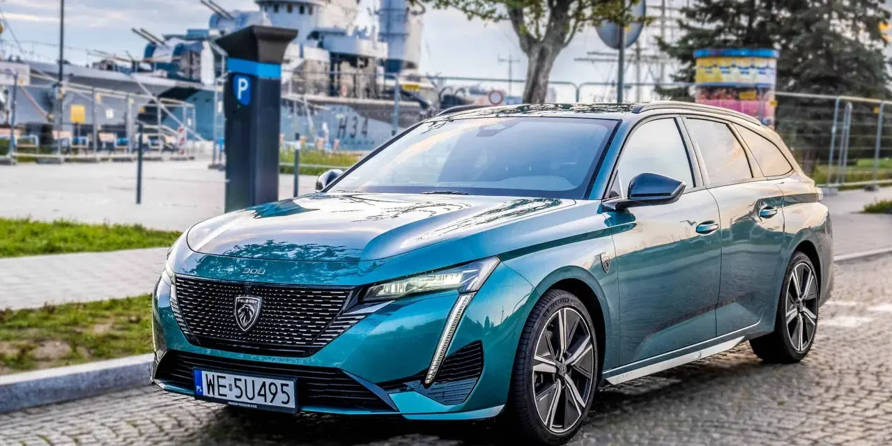 Turkusowe kombi Peugeot 308 zaparkowane na bruku, w tle okręt wojenny.