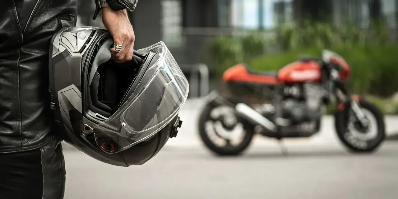 Motociclista in pelle tiene un casco, pronto per misurare la circonferenza della testa per un acquisto perfetto. Moto rossa sfocata sullo sfondo.
