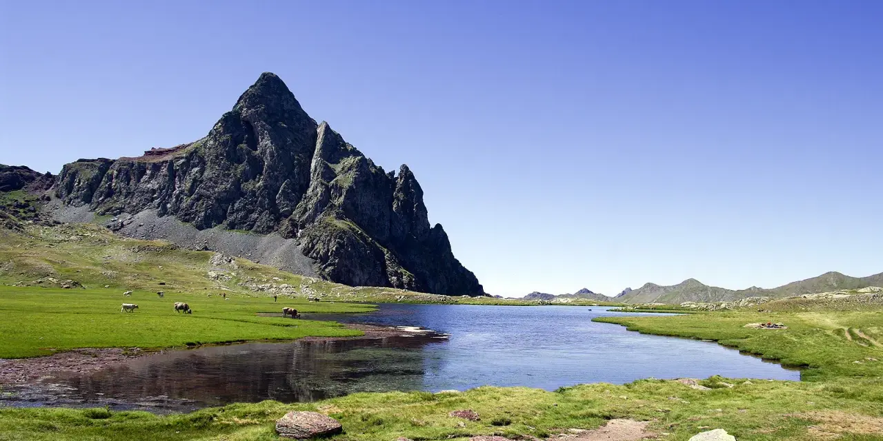Majestuoso pico rocoso se alza sobre un lago sereno y prados verdes en el valle de Tena, con vacas pastando tranquilamente.