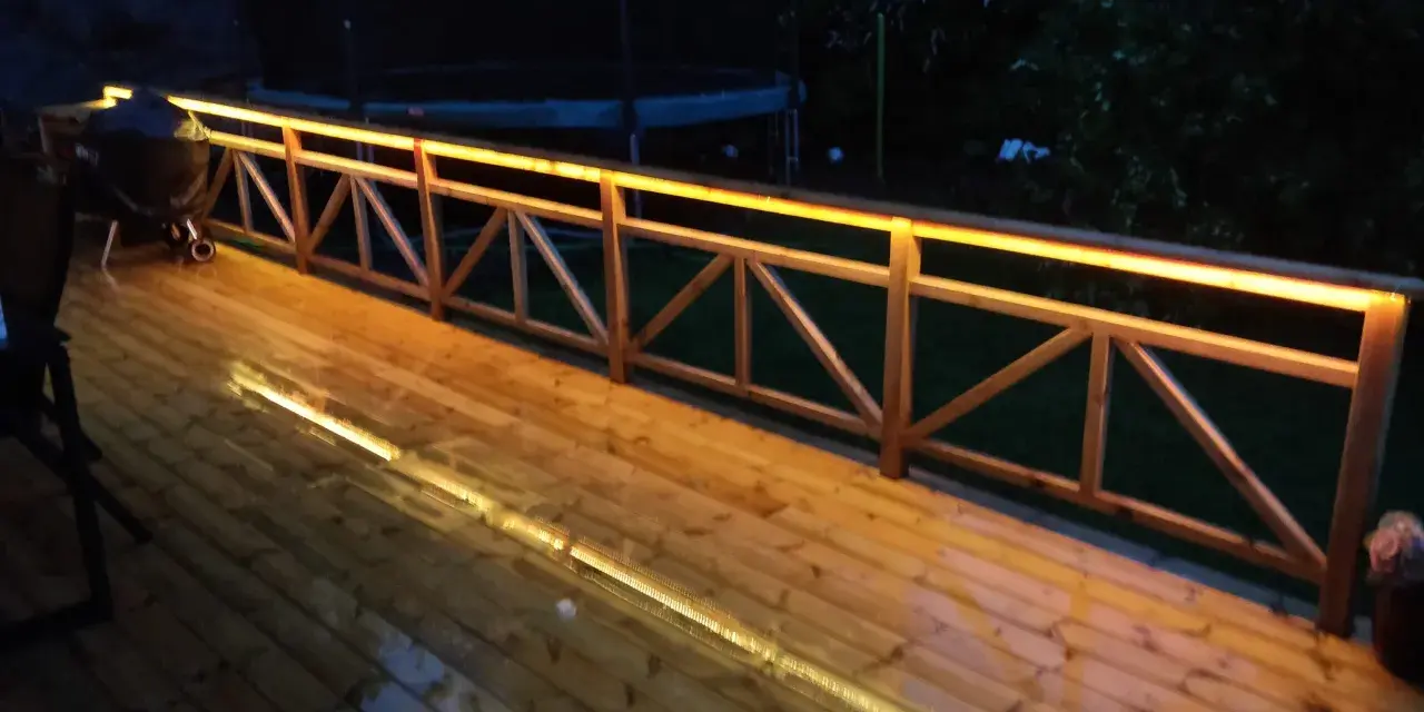 Träaltan med staket upplyst av varma decklights. En grill står på altanen.