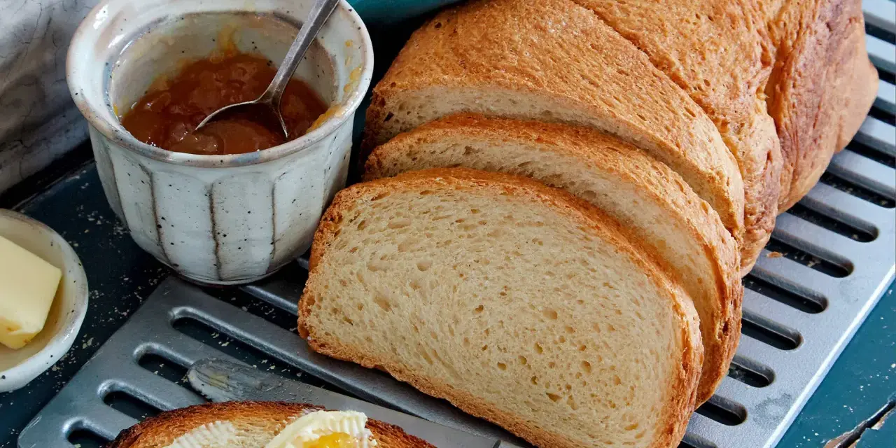Ein Laib **sauerteig Toastbrot** ist aufgeschnitten. Daneben stehen Marmelade, Butter und ein Messer mit einem Stück Brot, das bestrichen ist.