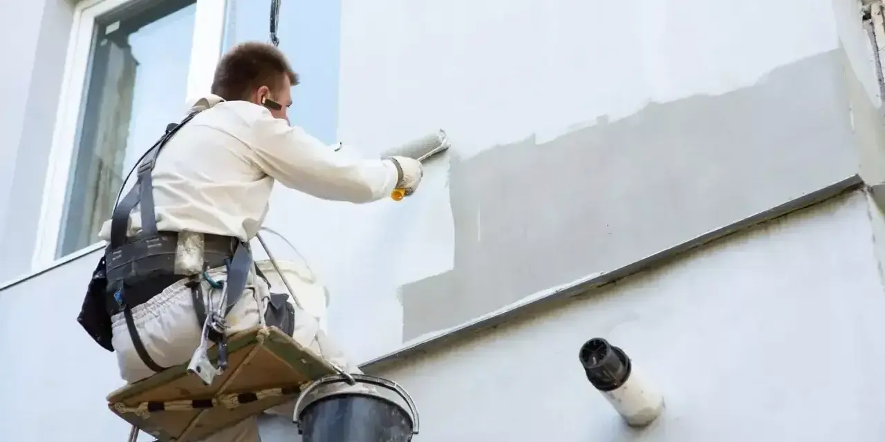 Malowanie elewacji cena: pracownik na platformie wiszącej maluje ścianę budynku wałkiem.