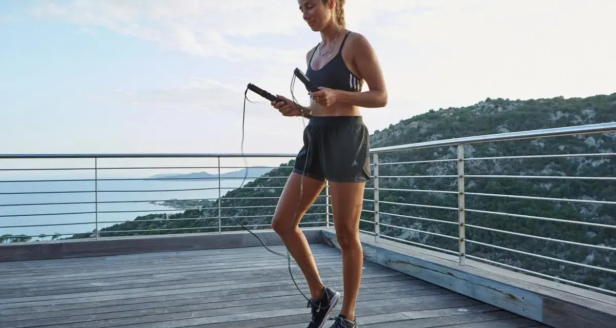 Femme en tenue de sport tenant une corde à sauter, prête pour un exercice en plein air avec vue sur la mer.