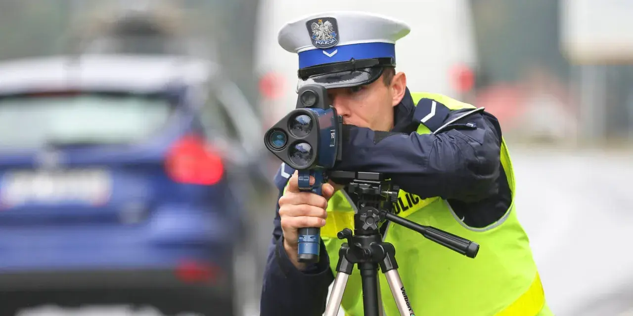 Policjant w kamizelce odblaskowej mierzy prędkość samochodu za pomocą radaru. Trwa dozór policyjny na drodze.