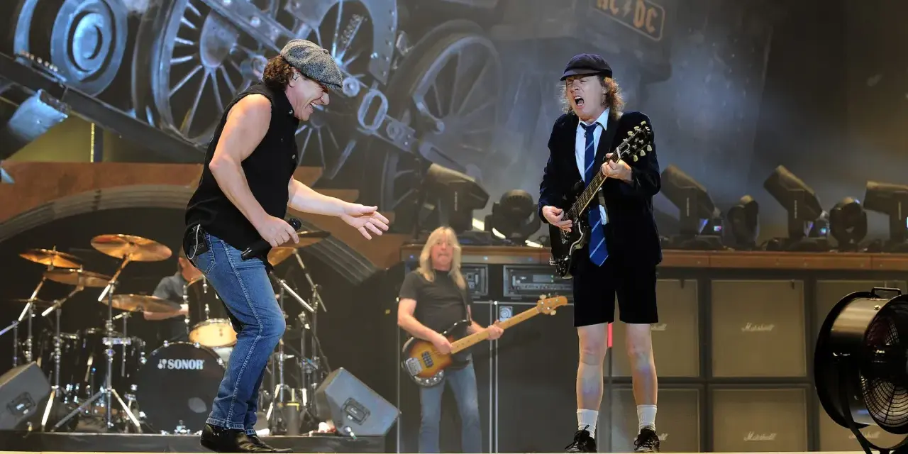 Brian Johnson singt, Angus Young spielt seine ACDC Gitarre. Im Hintergrund eine Dampflok.