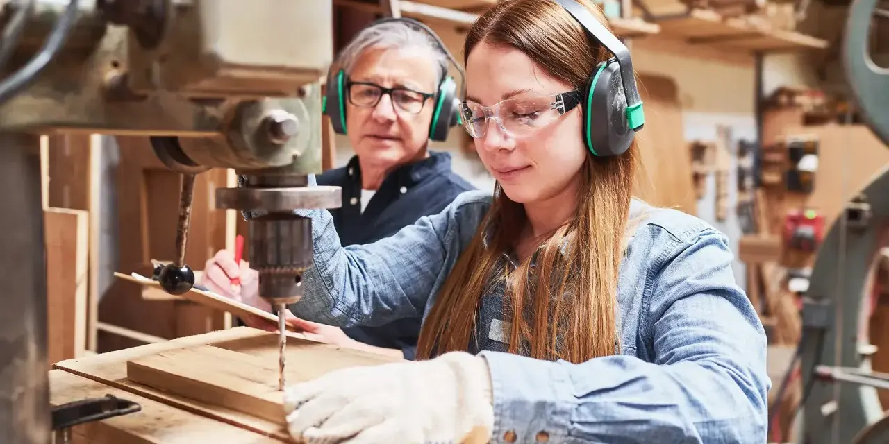 Junge Frau lernt an einer Bohrmaschine, während ein älterer Mann zusieht. Eine gute Ausbildung, welche Ausbildungen dauern 2 Jahre, ist hier vielleicht der Anfang.