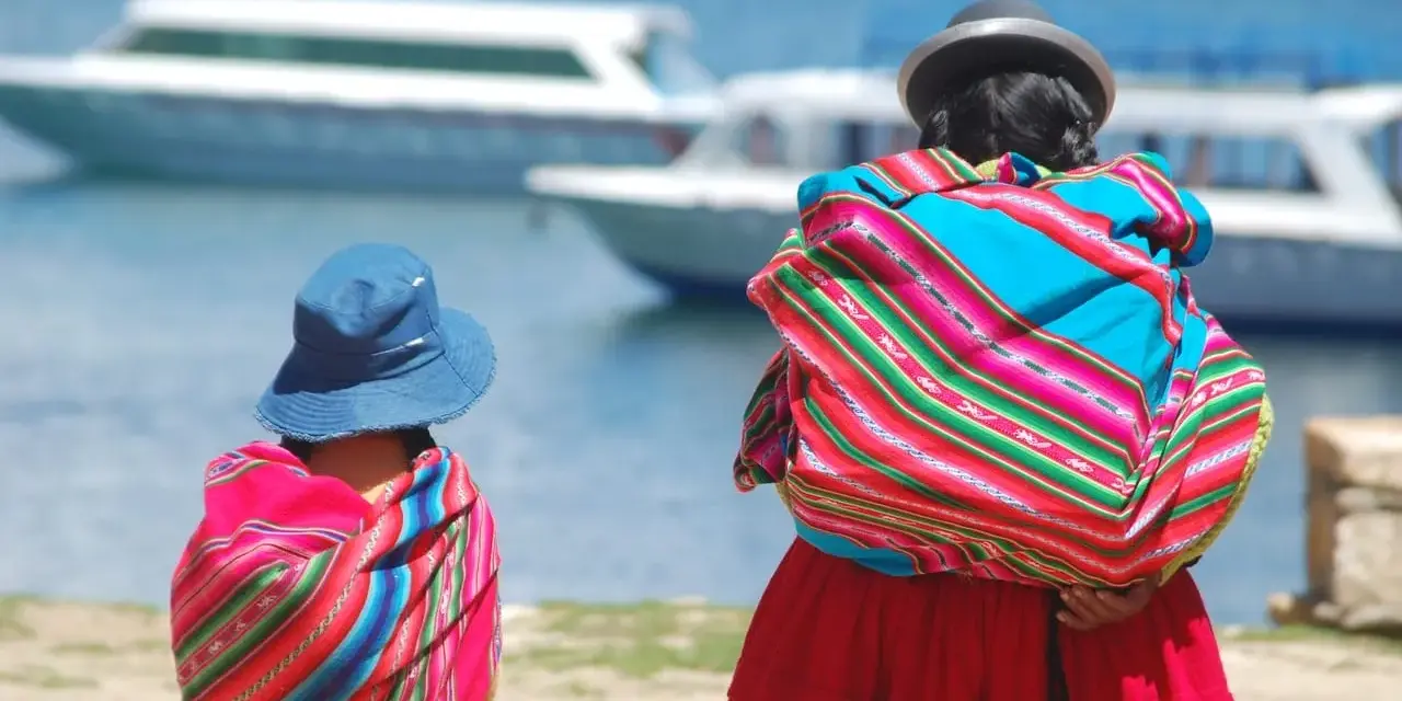 Zwei cholitas escaladoras, gekleidet in bunte Ponchos und Hüte, blicken auf Boote auf einem See.
