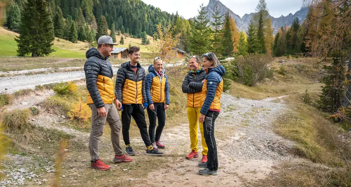Gruppo di escursionisti con giacche imbottite gialle e blu, pantaloni tecnici e scarponi. Perfetto esempio di come vestirsi in montagna per un'avventura autunnale.