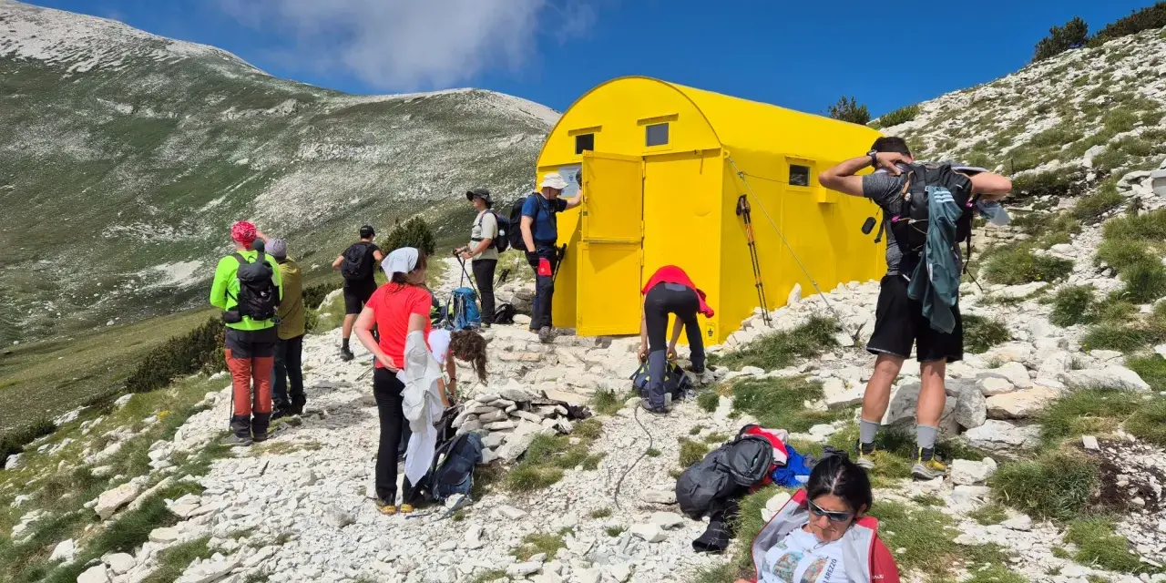 Gruppo di escursionisti vicino a un giallo blockhaus Majella, su un sentiero roccioso con montagne verdi sullo sfondo.