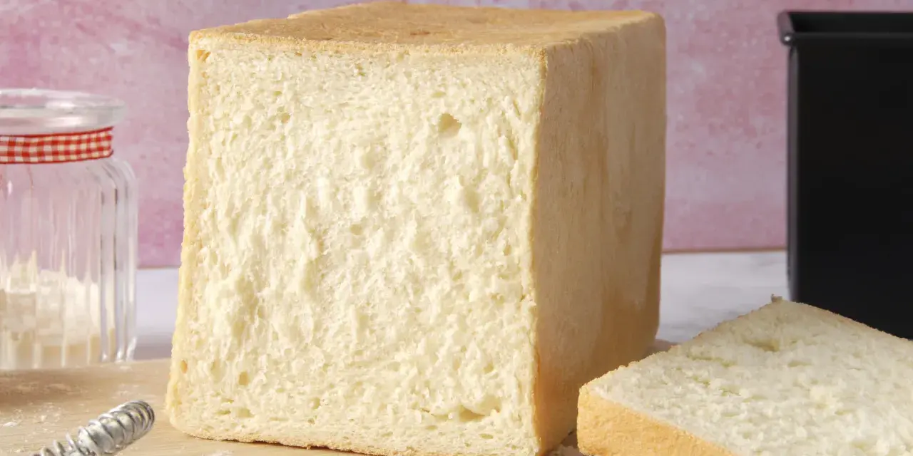 Un pain de mie moelleux, parfait pour une recette facile. Un pot de farine et un moule à pain sont à proximité.