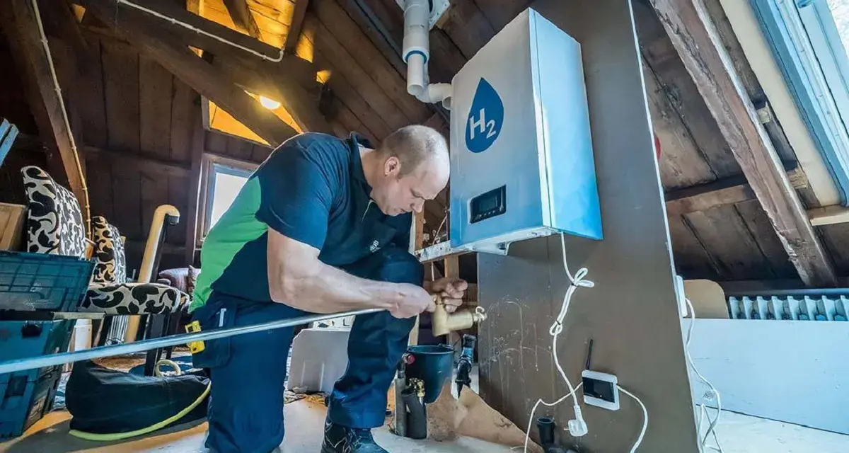 Installazione di caldaie a idrogeno in un sottotetto. Un tecnico collega tubi a un'unità bianca con il simbolo H2.