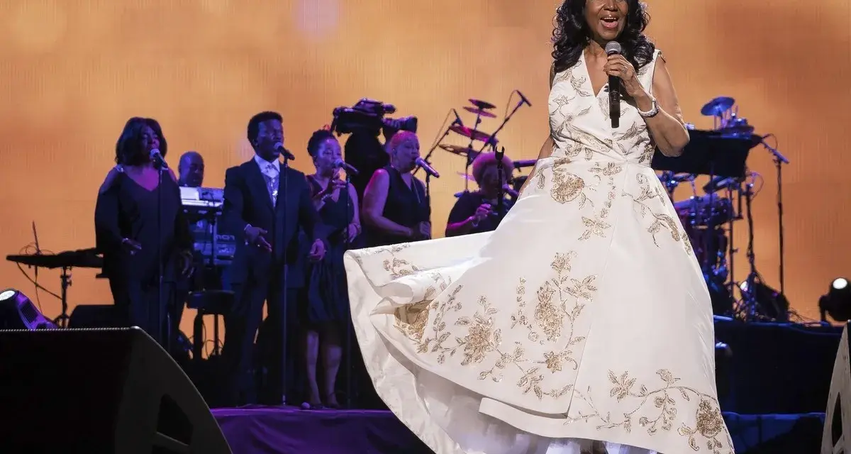 Aretha Franklin w białej sukni z haftem, śpiewa na scenie z chórem. Jej dzieci z pewnością byłyby dumne.