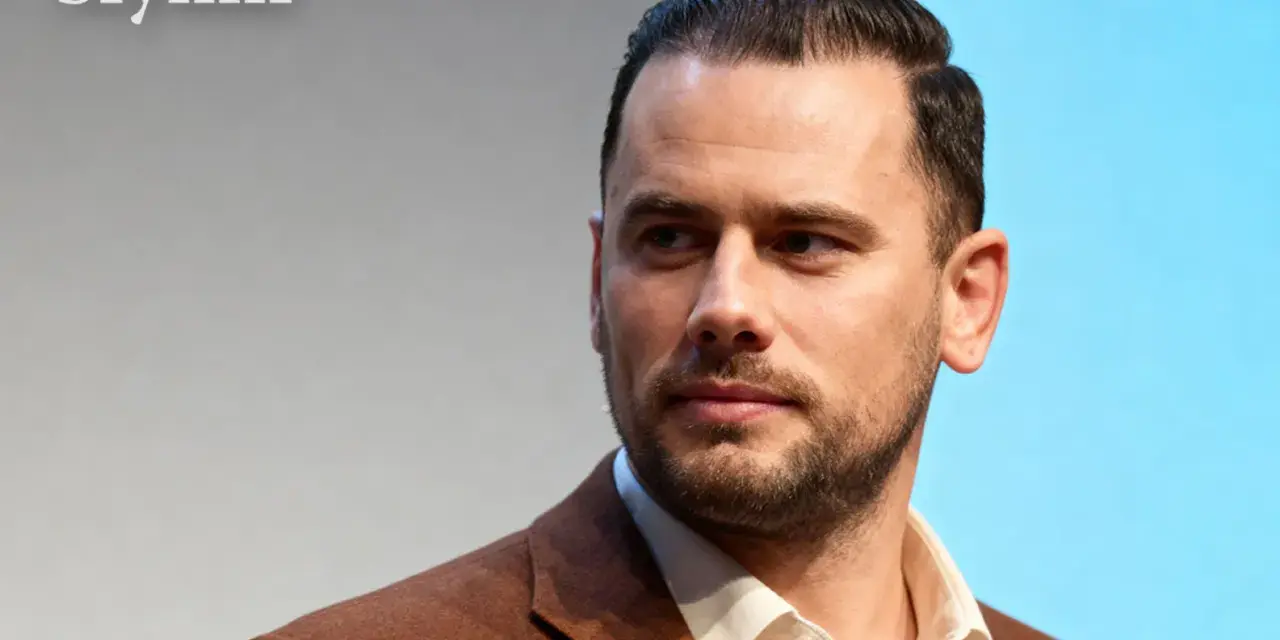 Grzegorz Collins, znany z programów telewizyjnych, w eleganckim brązowym garniturze. Jego wiek jest trudny do określenia, ale prezentuje się dojrzale.