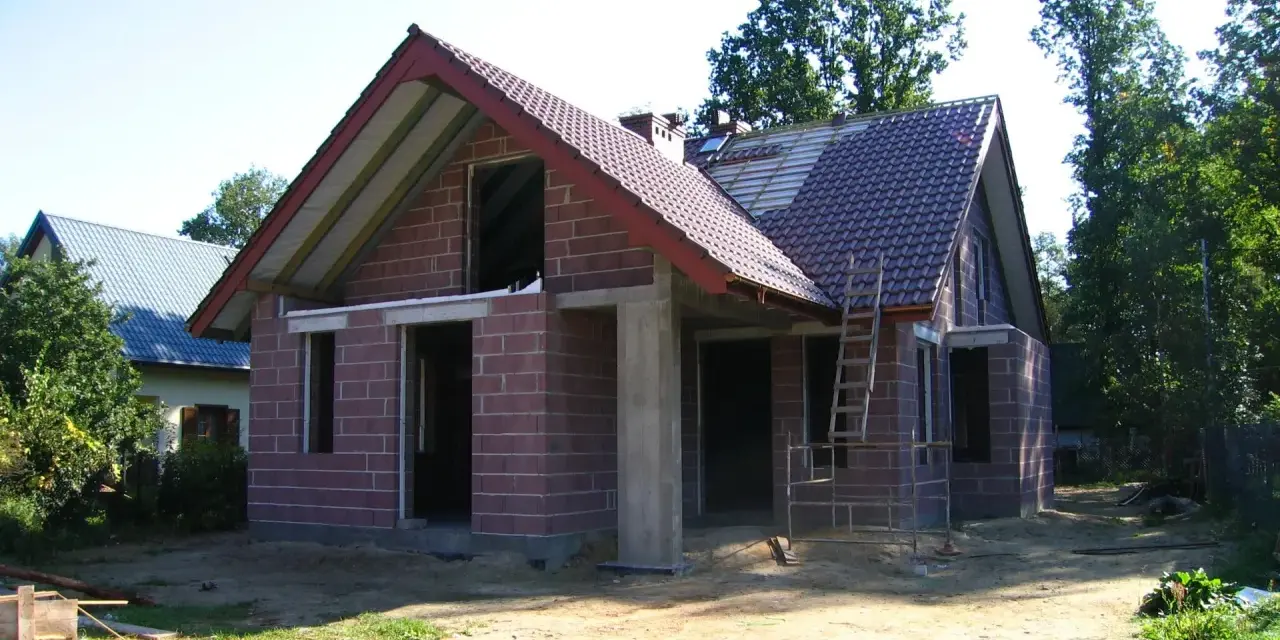 Dom w stanie surowym otwartym, z czerwonej cegły, z częściowo położonym dachem. Wokół plac budowy.