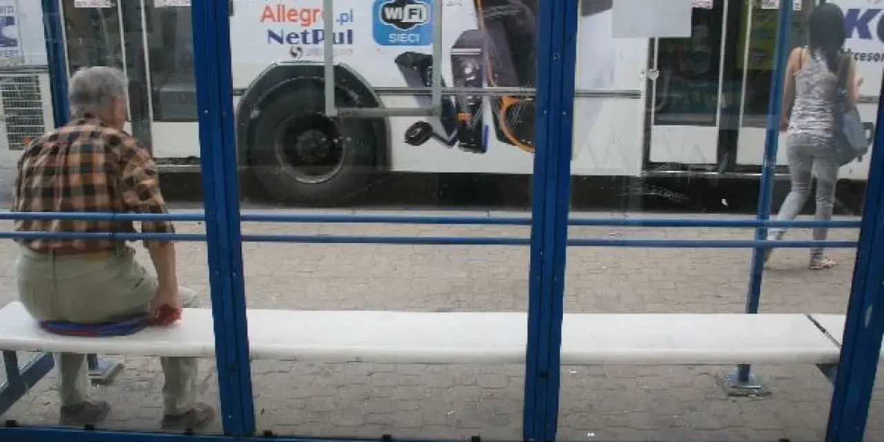 Mężczyzna czeka na przystanku na autobus 102. Rozkład jazdy jest niewidoczny, ale widać autobus z reklamą Allegro.