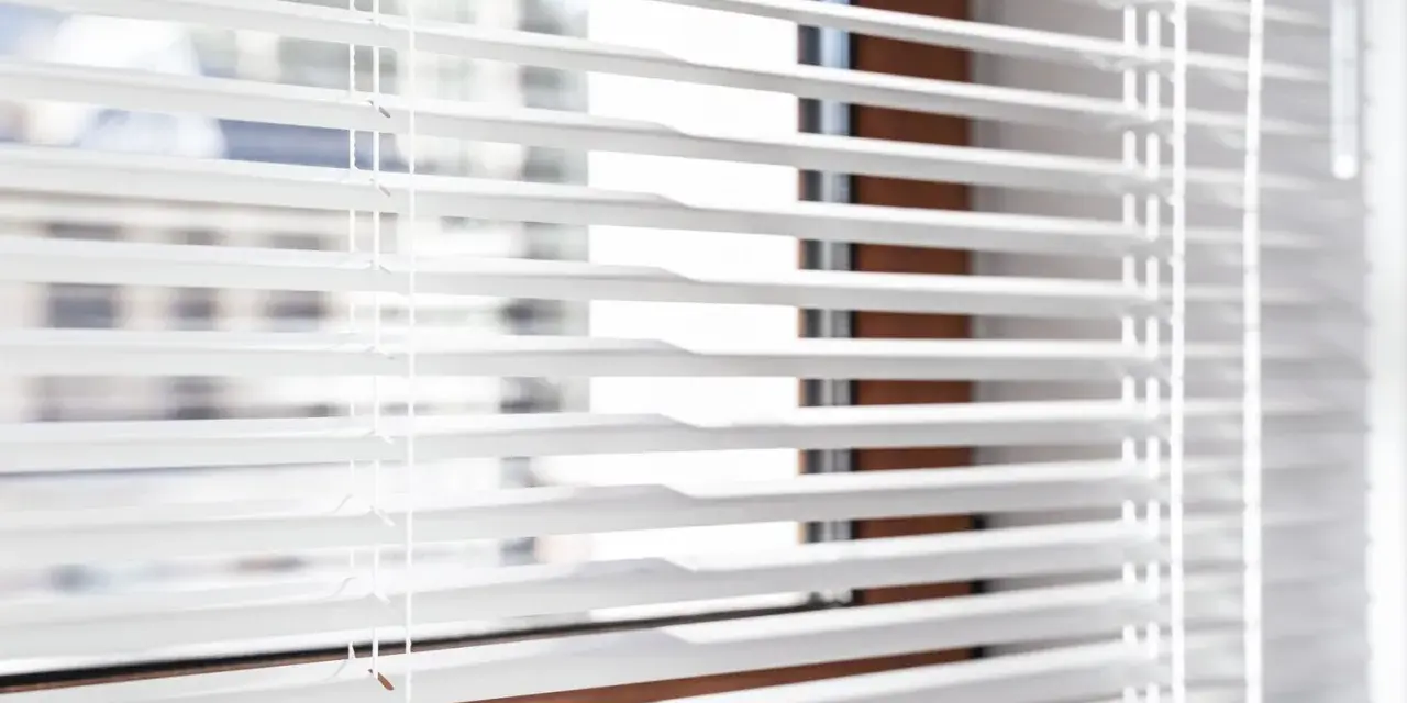 Close-up of white horizontal blinds with visible cords, showing the slats and a hint of the window frame.