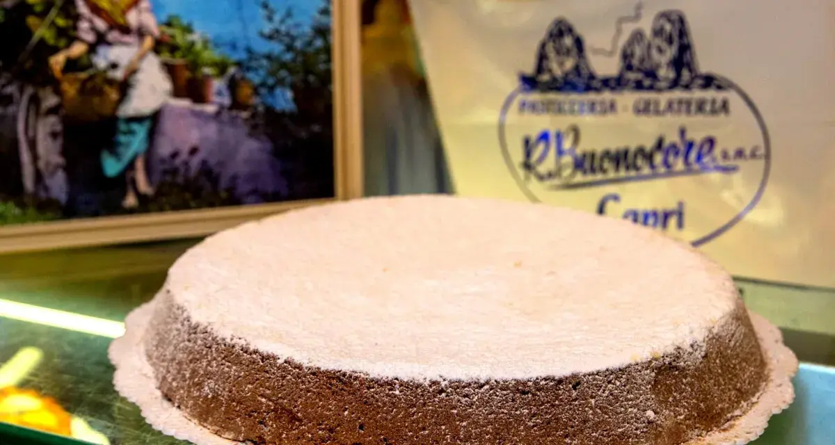 A Torta Caprese dusted with powdered sugar sits on a glass counter, with a painting and shop logo in the background.