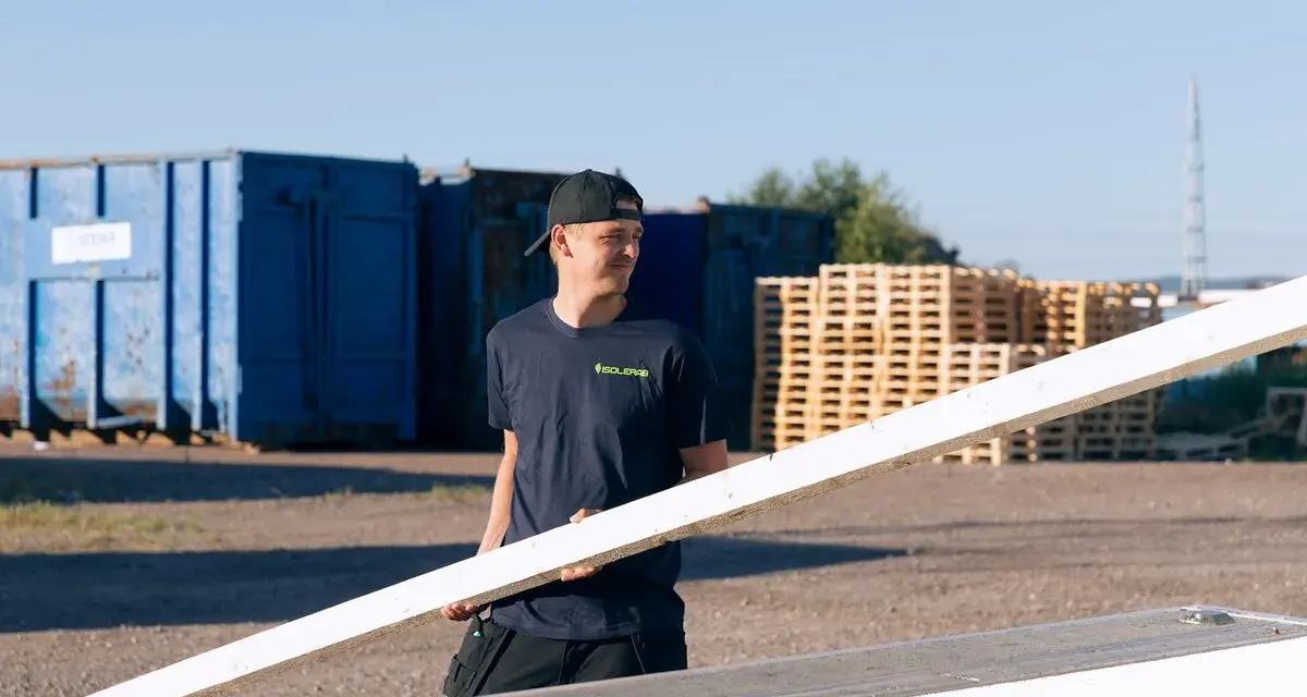 Man i mörkblå t-shirt med "ISOLERAB" logga bär en träskiva. Bakom honom syns blå containrar och träpallar.