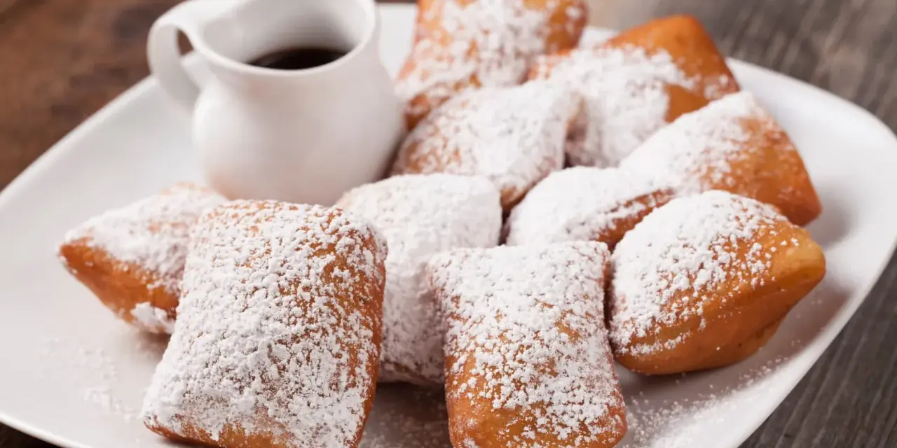 Une assiette de beignets saupoudrés de sucre glace, parfaits pour une recette pâte à beignets salés. Un petit pichet de sauce est à côté.