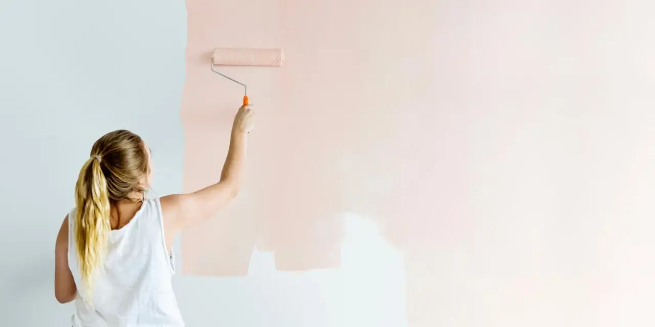 Frau streicht mit einer Malerrolle eine Wand in einem zarten Rosaton. Die Wohnung bekommt einen neuen Anstrich.