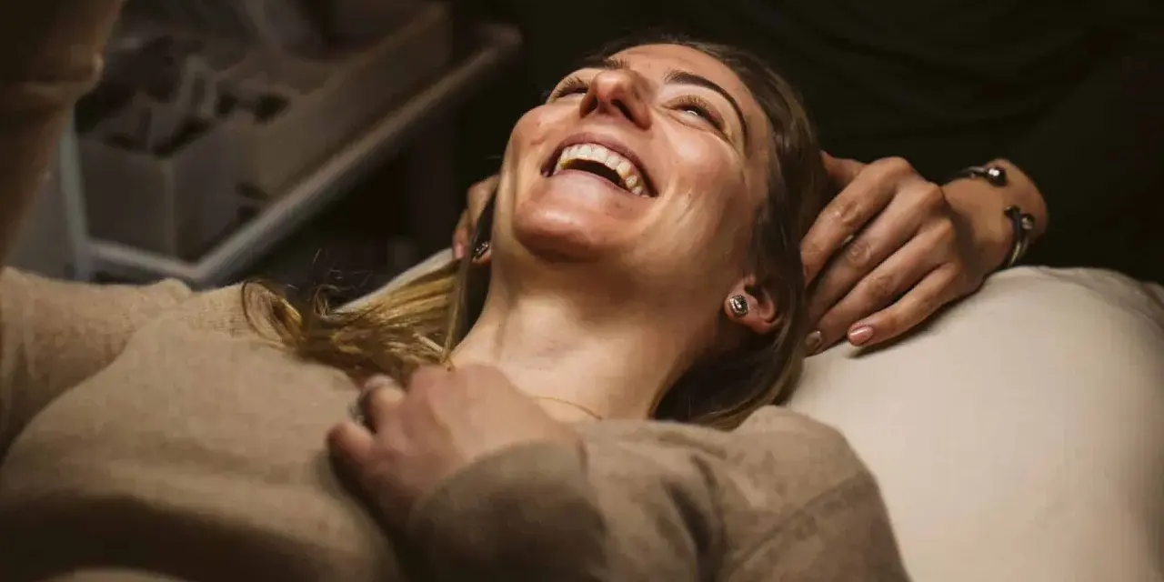 A woman laughs joyfully during a holistic facial treatment, her head tilted back as hands gently support her.