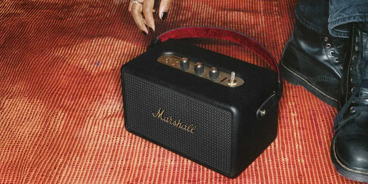 En svart Marshall Kilburn-högtalare på en röd, randig matta. En hand med ring och nagellack sträcker sig mot den.