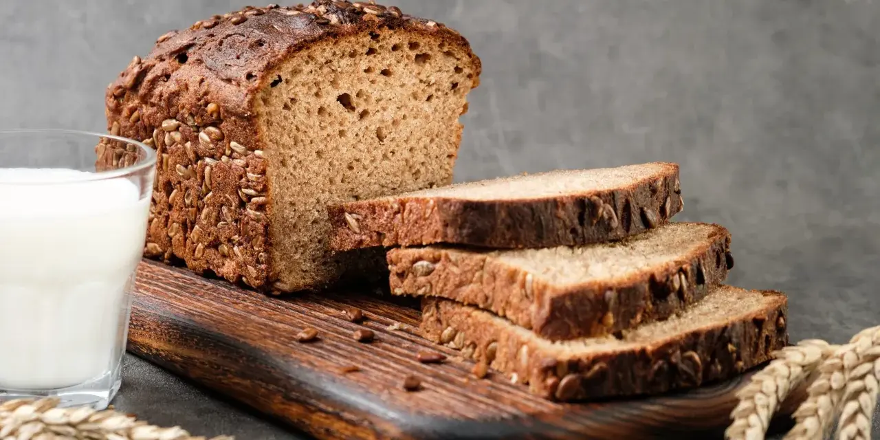 Chleb proteinowy z ziarnami, pokrojony na kromki, obok szklanka mleka i kłosy zboża. Idealny na zdrowe śniadanie.