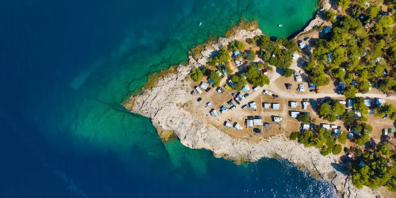 Widok z lotu ptaka na kemping Riviera Makarska. Kamieniste wybrzeże, turkusowa woda i zielone drzewa otaczają liczne przyczepy kempingowe i samochody.