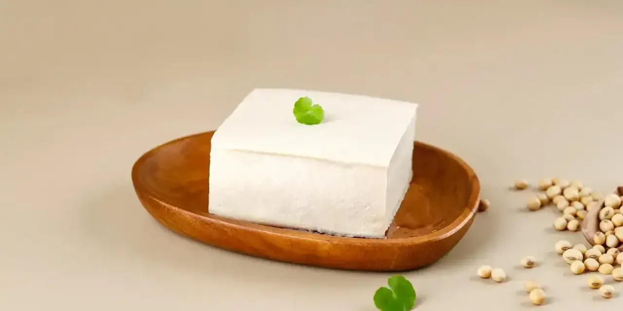 Kostka białego tofu na drewnianej miseczce, ozdobiona listkiem. Obok garść ziaren soi. To właśnie co to tofu – produkt z soi.