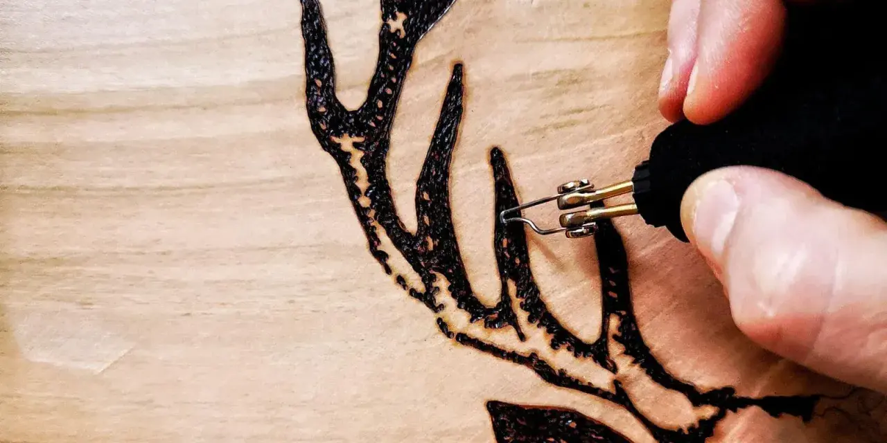 Dettaglio di un disegno a pirografia su legno: una mano guida le punte pirografo per creare le corna di un cervo.