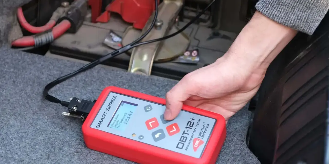 Ręka trzyma tester akumulatora DBT-12+, pokazujący 12.14V. To prosty sposób, jak sprawdzić akumulator w samochodzie.