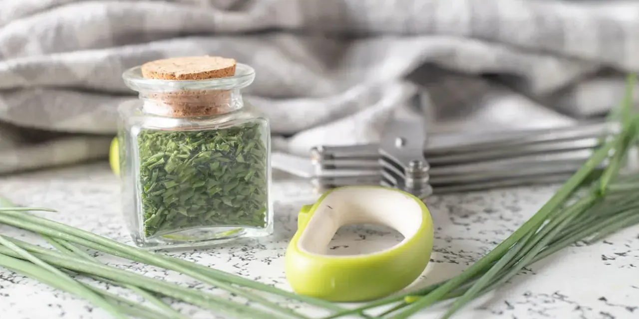 A jar filled with dried chives, showcasing how to dry chives for long-term storage. Fresh chives and kitchen tools are nearby.