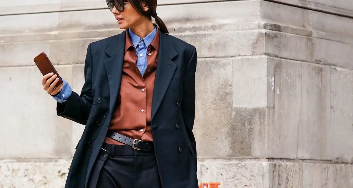 Mujer con estilo combinando traje negro con camisa marrón y azul, gafas de sol y móvil en mano.