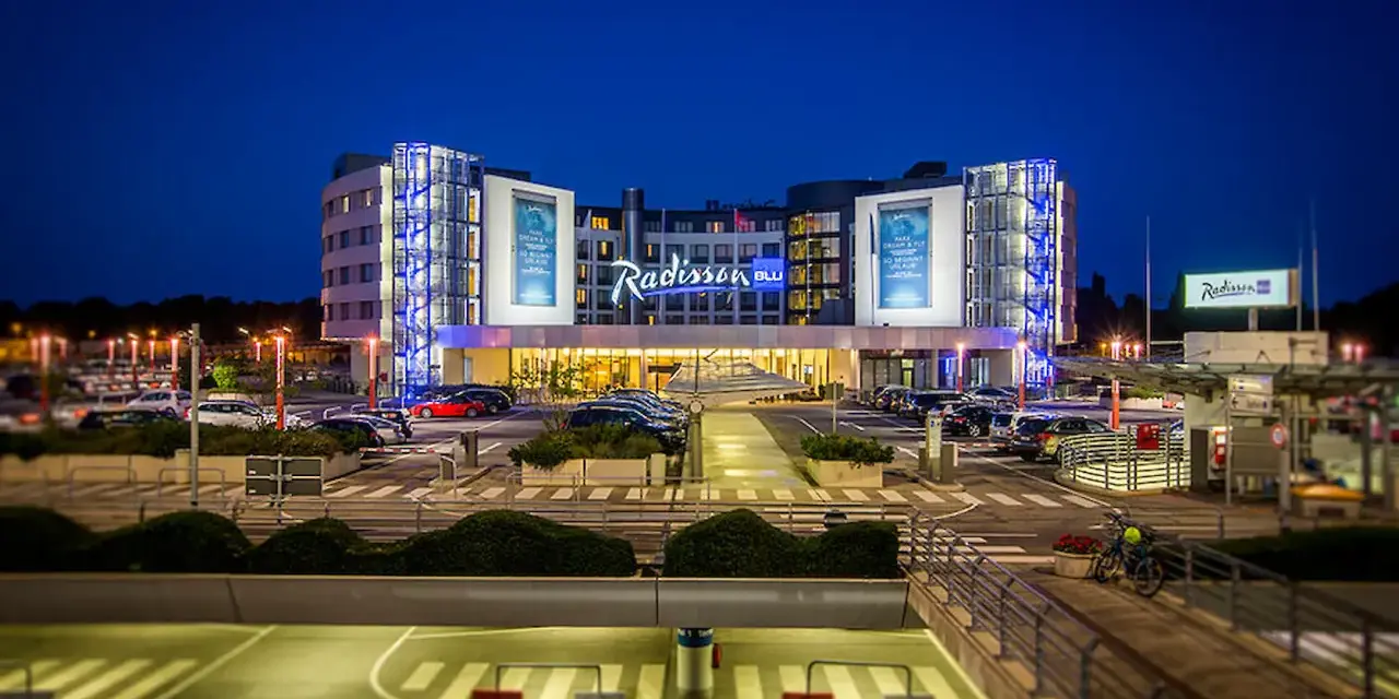 Nowoczesny hotel Radisson Blu Hamburg Airport nocą, z podświetlanym logo i parkingiem pełnym samochodów.