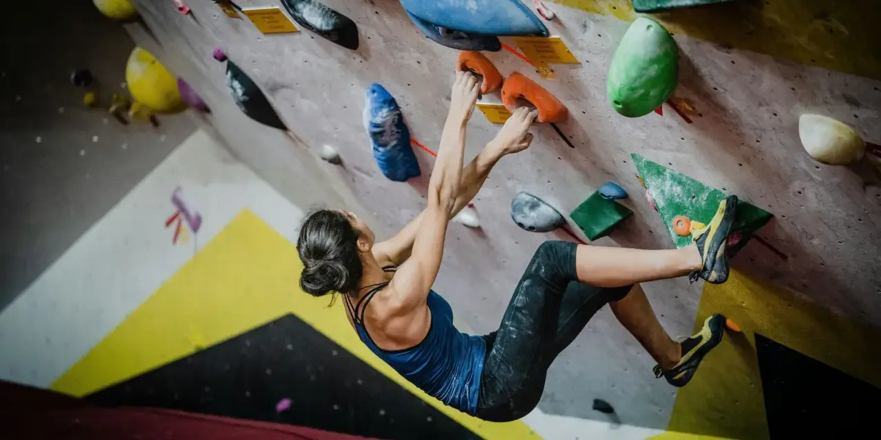 Kobieta w niebieskiej koszulce wykonuje trudny ruch podczas wspinaczki sportowej na kolorowej ściance.