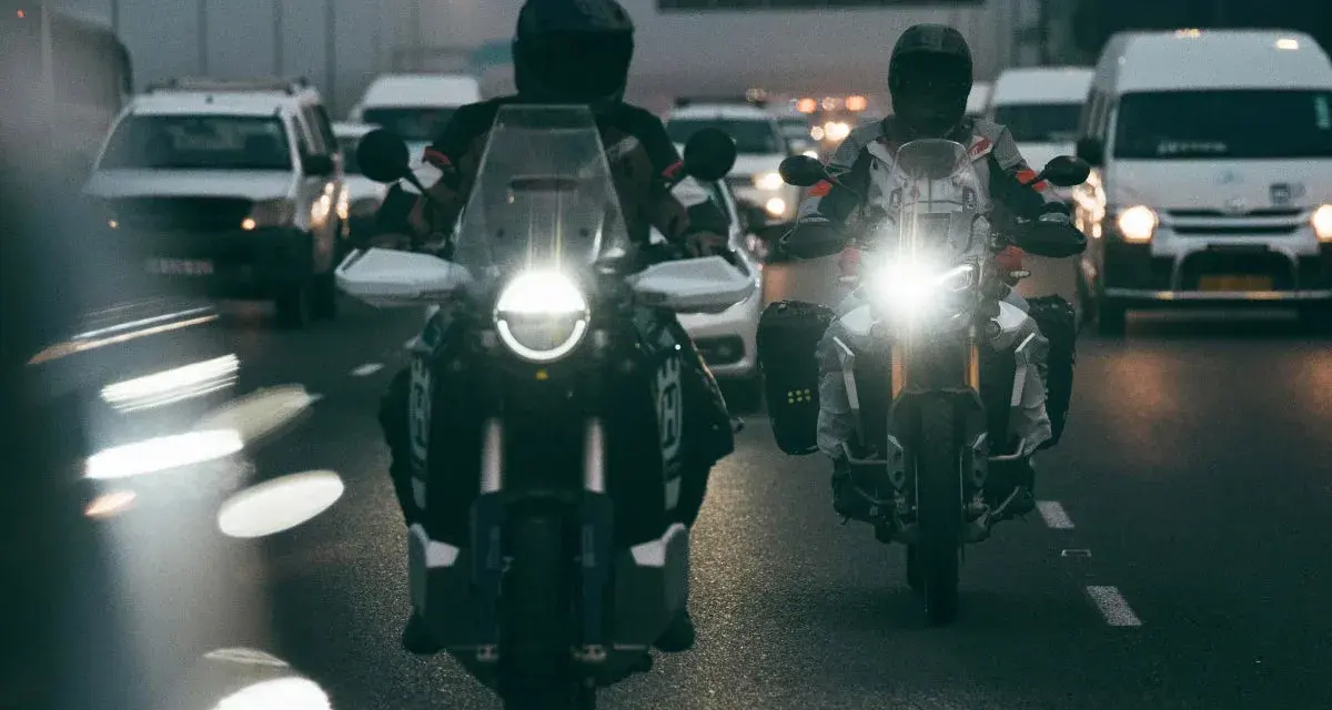 Dwóch motocyklistów w kaskach jedzie w deszczu. Ich motocykle mają włączone światła, a droga polna jest mokra i błyszcząca.
