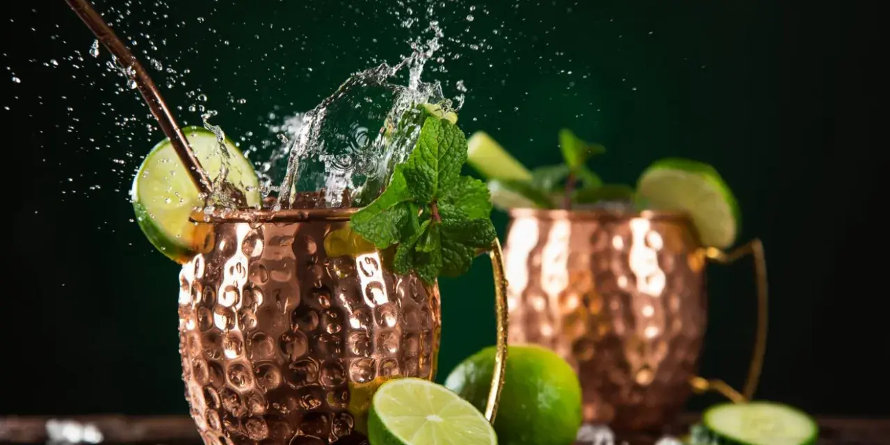 Dwa miedziane kubki z orzeźwiającym Moscow Mule, ozdobione limonką i miętą, z bryzgami wody i kostkami lodu.