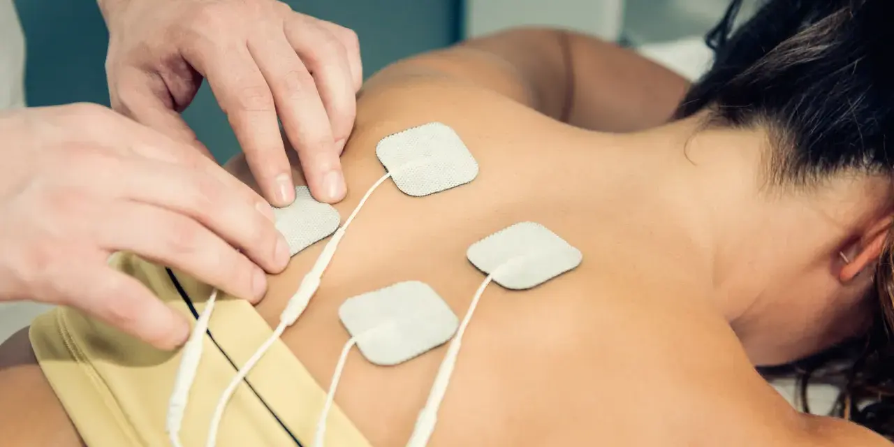 Fizjoterapeuta umieszcza elektrody na plecach pacjentki, przygotowując do zabiegu TENS. Należy pamiętać o przeciwwskazaniach do jego stosowania.