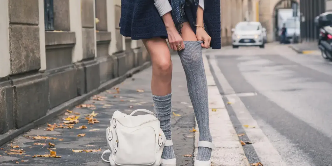 Stylizacja z długimi skarpetkami damskimi, biały plecak i jesienne liście na chodniku.