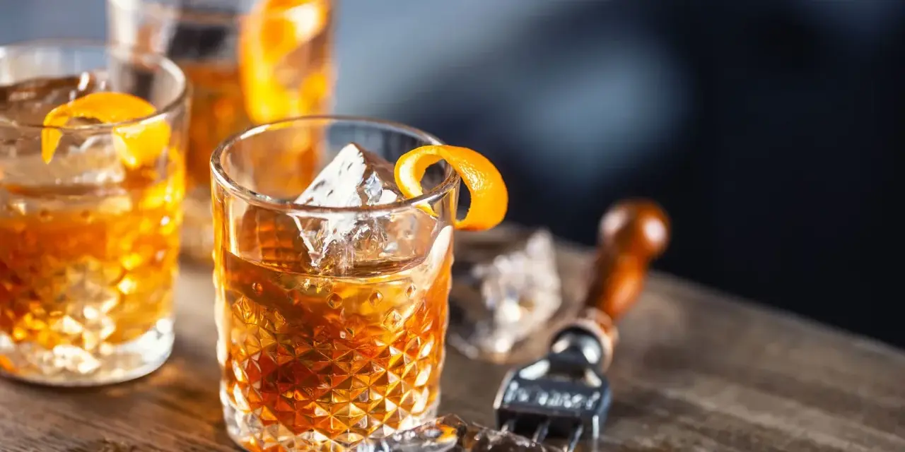 Tre old fashioned-drinkar med is och apelsinskal på ett träbord. En cocktailgaffel syns i bakgrunden.