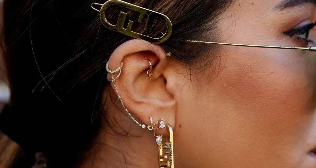 Ucho ozdobione wieloma kolczykami, w tym forward helix piercing, z błyszczącymi dodatkami i złotą spinką z logo.