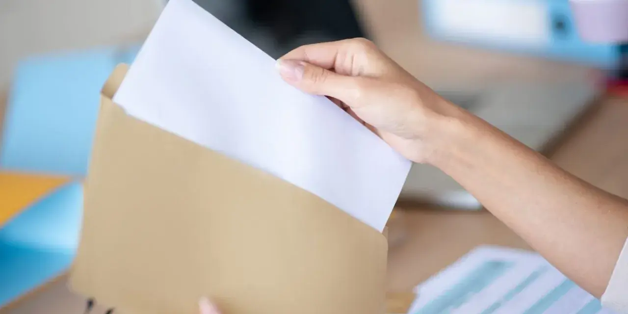 Dłoń wkłada biały papier do brązowej koperty. Wzór adresowania koperty A4 jest gotowy do wypełnienia.